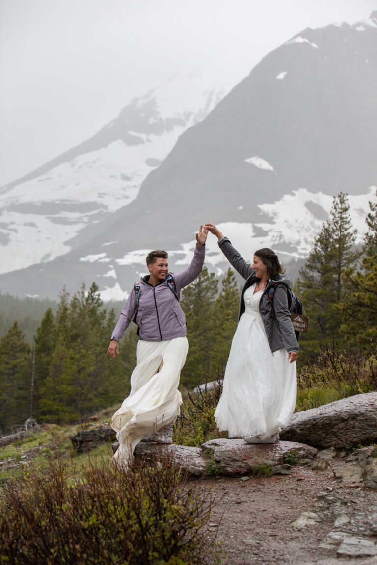 Two brides stand on a rock dancing in the rain on their leave no trace elopement day.