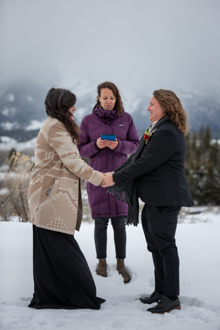 Two brides stand facing each other while their officiant reads the ceremony script on their leave no trace elopement day.