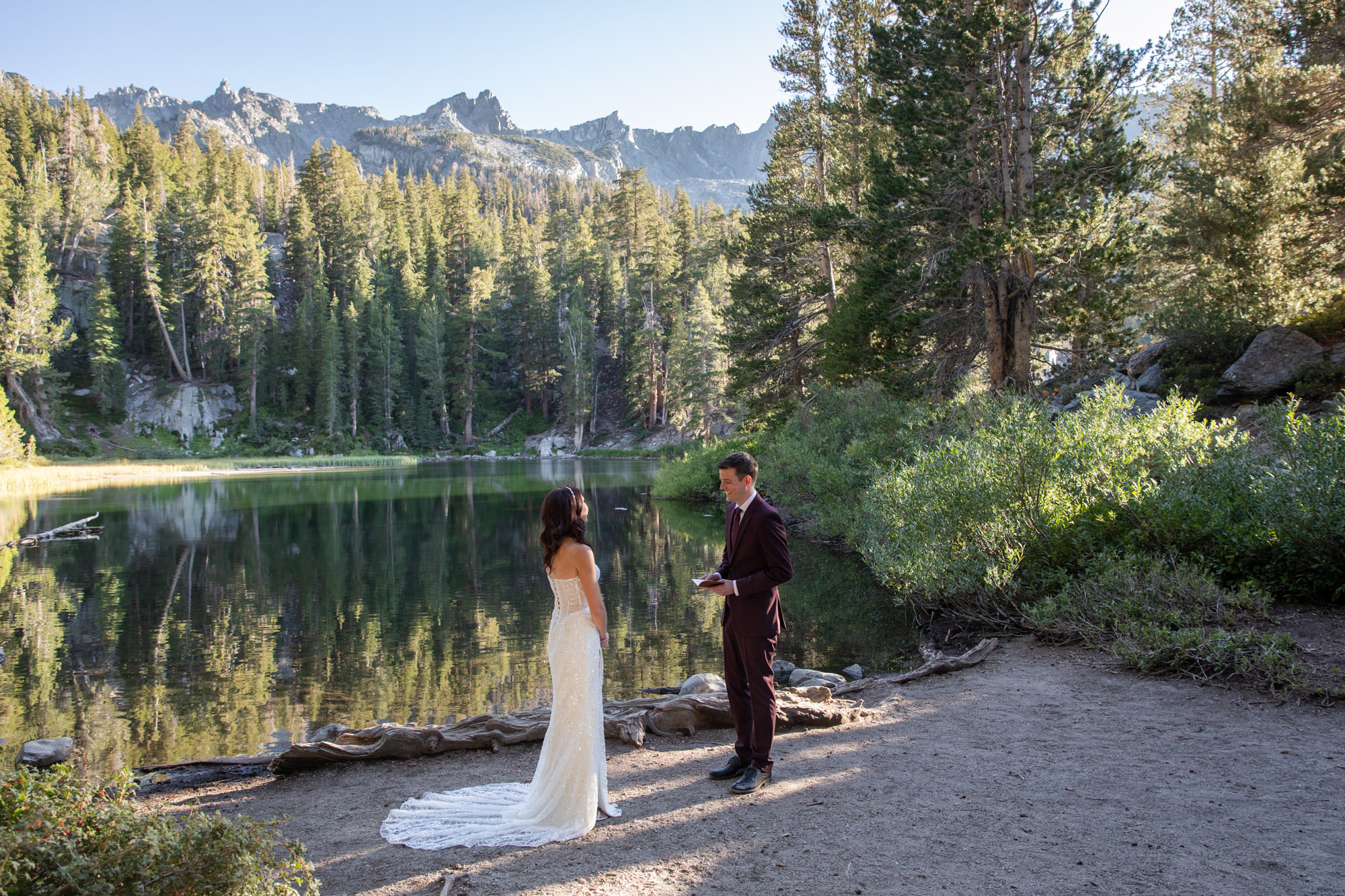 A bride wearing a white lace dress stands facing her groom who wears a burgundy suit standing next to a lake on their Mammoth Lakes elopement day.