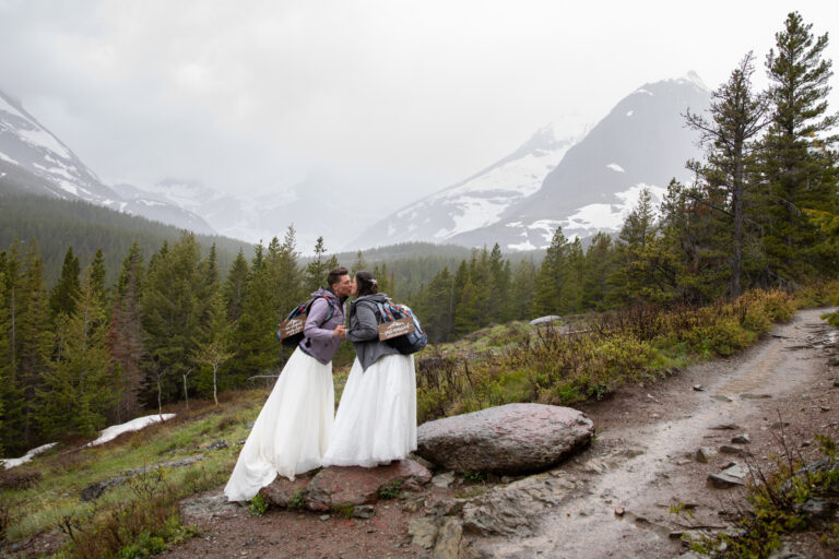 Two brides stand in the rain holding hands and kissing during their Many Glacier Elopement day.