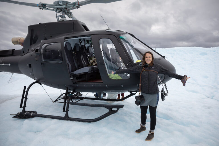 Becky stands next to a helicopter in Alaska wearing two of her must have camera accessories, her Rose Anvil Harness with her Canon 5d Mark IV