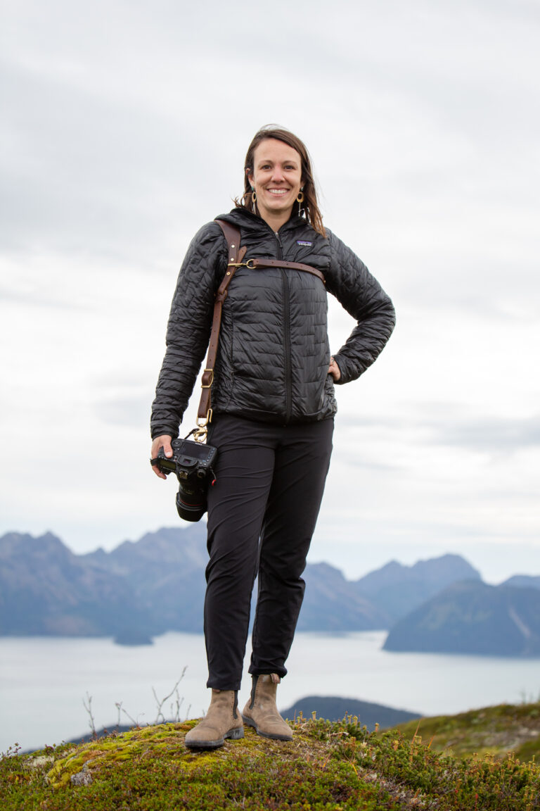 Becky stands on a mountain top in Alaska, wearing two of her must have camera accessories, her Rose Anvil camera harness and her Canon 5d Mark IV