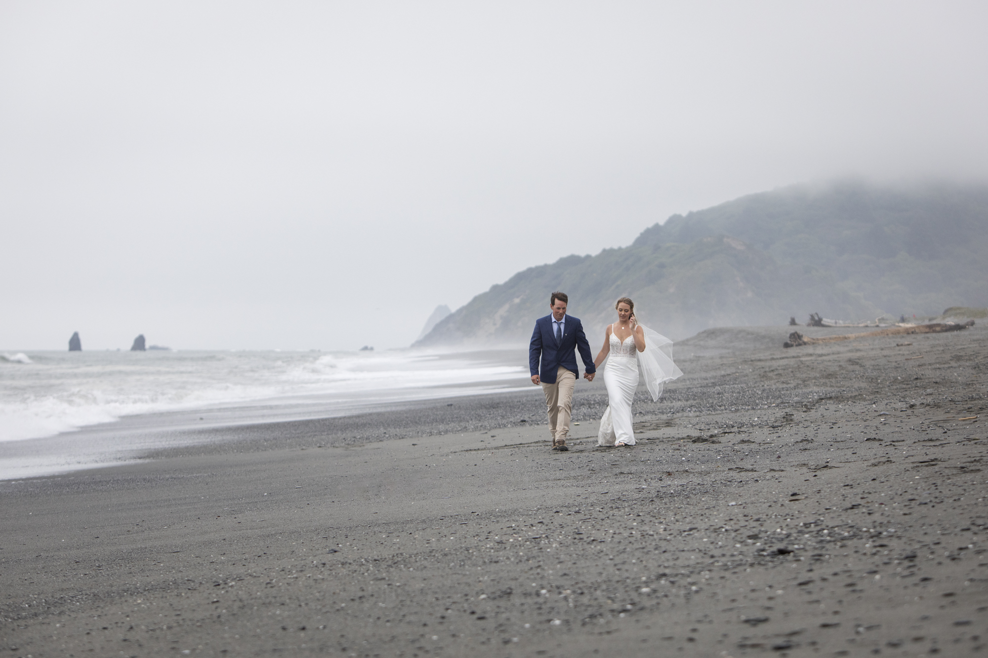 A bride and groom walk hand in hand on their Oregon Coast elopement day, as the waves break along the shore.
