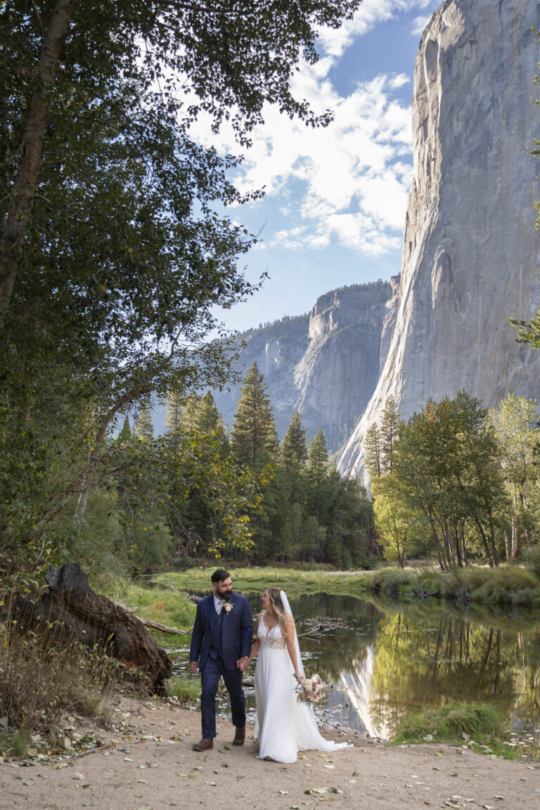 10 Breathtaking Places in California to Elope | Scenic Vows A bride and groom walk on a beach next to a river, smiling at each other on their wedding day in Yosemite, one of the best places in California to elope.
