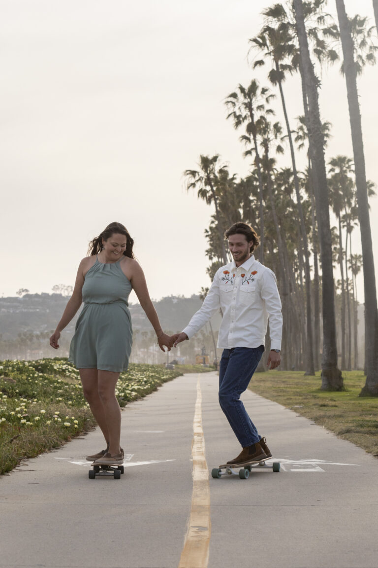 10 Breathtaking Places in California to Elope | Scenic Vows A woman in a green skirt rides a skateboard and holds hands with a man in a white shirt with orange flowers also riding a skateboard down a paved path.