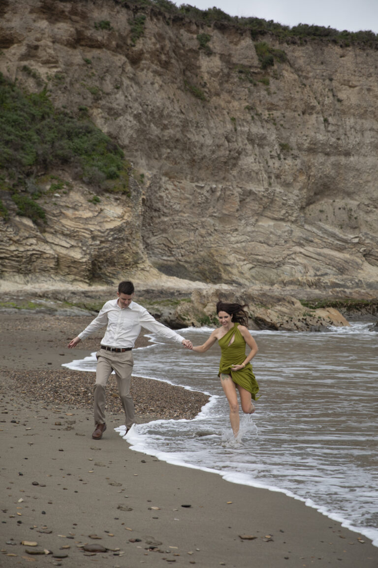 10 Breathtaking Places in California to Elope | Scenic Vows A woman in a green dress holds hands with a man in a white shirt and khaki pants as they run in the waves along the beach.