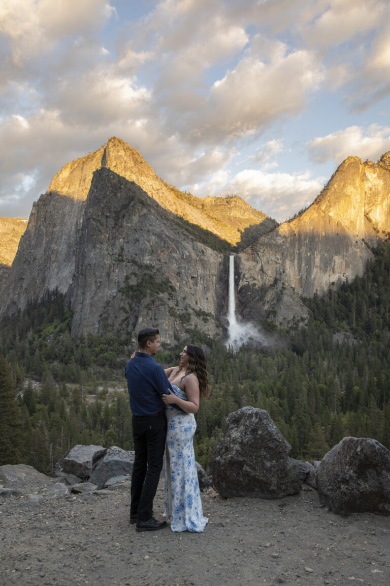 10 Breathtaking Places in California to Elope | Scenic Vows A bride and groom stand on a trail embracing with a waterfall and sunset in the background behind them.