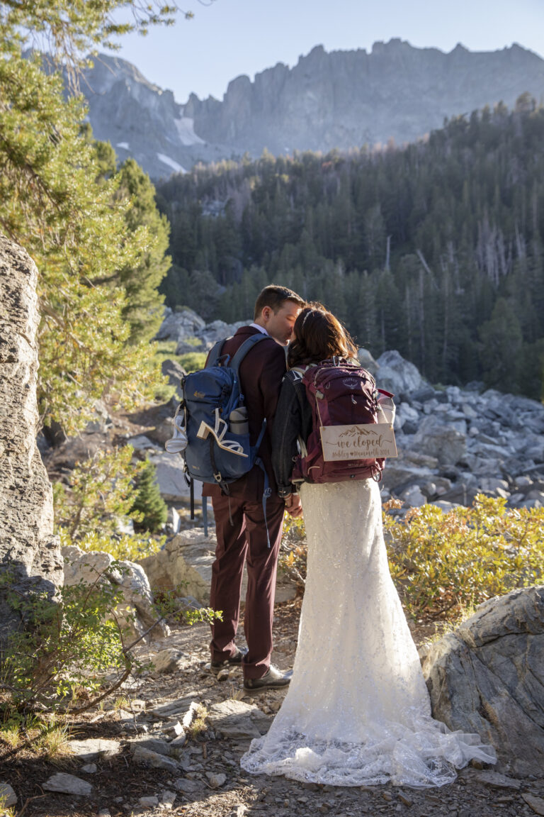10 Breathtaking Places in California to Elope | Scenic Vows A bride and groom wearing backpacks with "we eloped" signs, kiss while standing in the sun and mountains surround them.