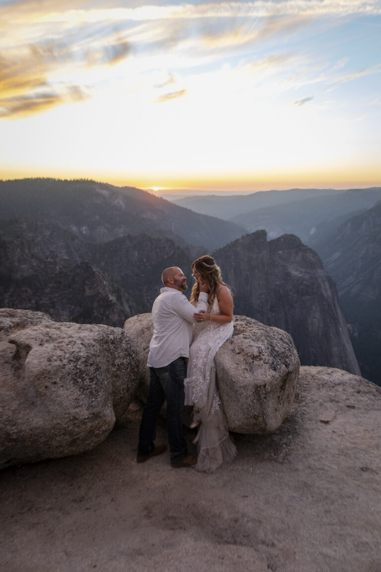 10 Breathtaking Places in California to Elope | Scenic Vows A bride sits on a rock while her groom holds her face and pulls her in for a kiss, as the sunsets behind them over the mountains.