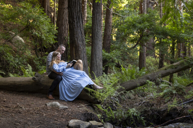 10 Breathtaking Places in California to Elope | Scenic Vows A man sits on a log while a woman in a blue dress leans into his chest reading a book aloud.