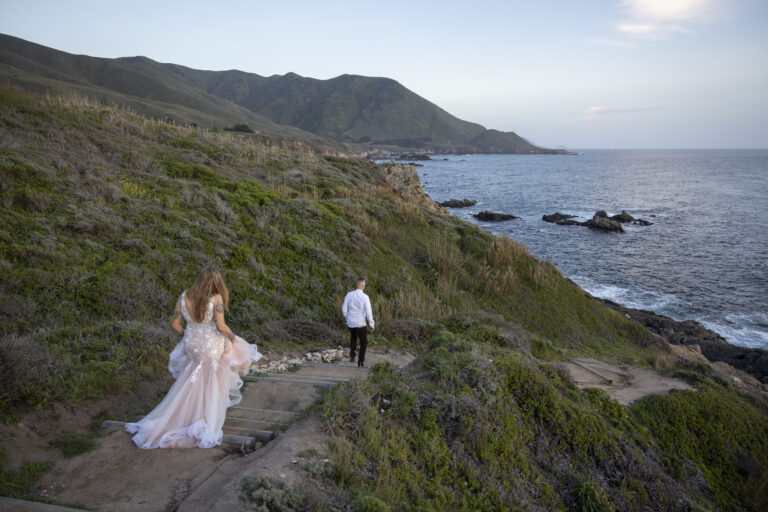 10 Breathtaking Places in California to Elope | Scenic Vows A man in a white shirt and black pants walk down dirt stairs towards the ocean as a woman in a wedding dress follows behind him.
