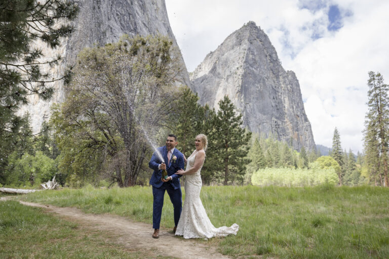 10 Breathtaking Places in California to Elope | Scenic Vows A bride and groom spray a bottle of champagne while standing on a trail in the middle of a meadow surrounded by mountains.