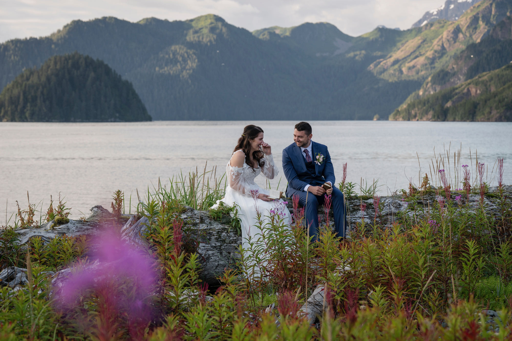 A bride laughs as her groom smiles at her while they sit on a log together with bright purple flowers surrounding them and the ocean behind them.