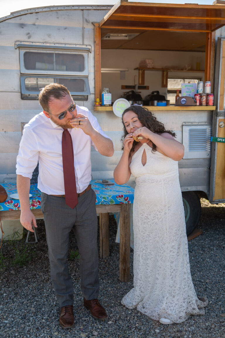 A bride and groom each take a bite of a taco in their hands.