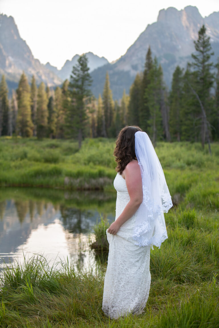 A bride stands next to a body of water, looking at the mountains in the background.