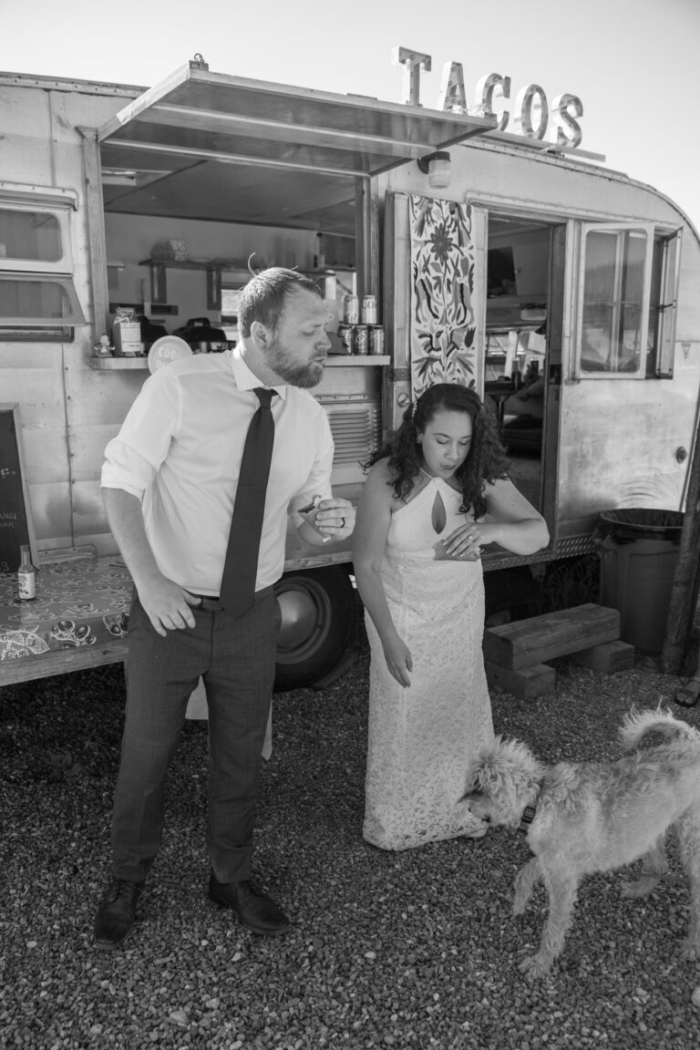 A couple stand in front of a taco truck with tacos in their hands and a dog at their feet.