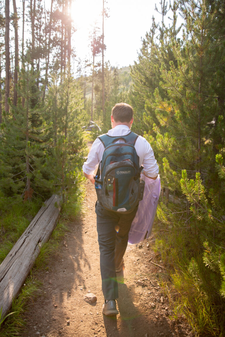 A man wearing a backpack and carrying a bag walks along a dirt path with trees all around him.