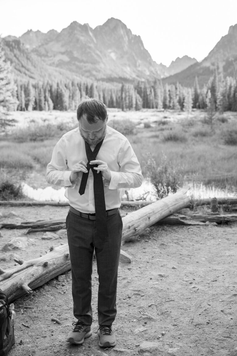 A man stands next to a log in the woods, tying his tie for his Stanley Idaho elopement ceremony.