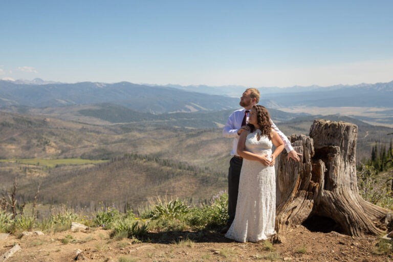 A groom stands behind his bride, leaning against a tree stump as she stands in front of him with her hands on her waist.