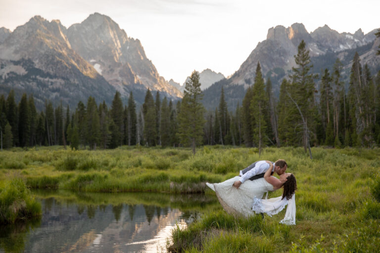 A groom dips and kisses his bride next to a creek in Stanley Idaho on their elopement day.