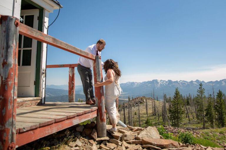 A groom helps with bride up onto the wooden deck of a fire tower lookout on their Stanley Idaho elopement day