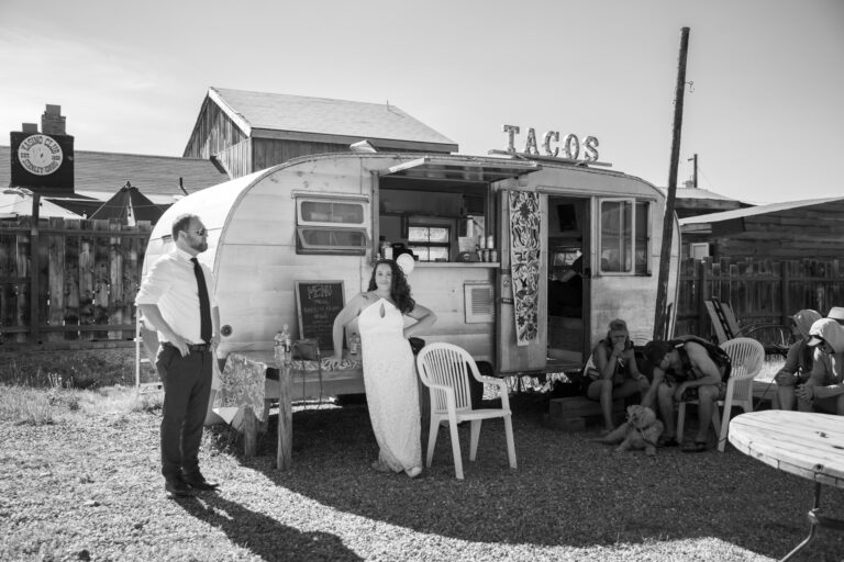 A bride and groom stand in front of a taco truck in Stanley Idaho while some locals sit to the right petting a dog.