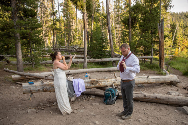 A bride does her makeup in a hand held mirror while a groom ties a tie in the woods.