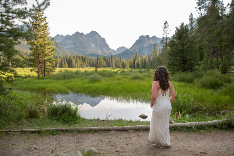 A bride stands looking out over a small lake with mountains in the background.