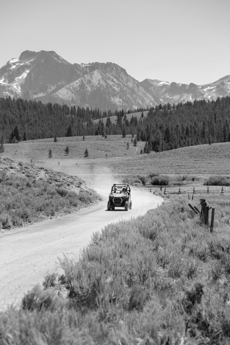 A black and white image shows a side-by-side driving down a dirt road with the Sawtooth Mountain range in the background.