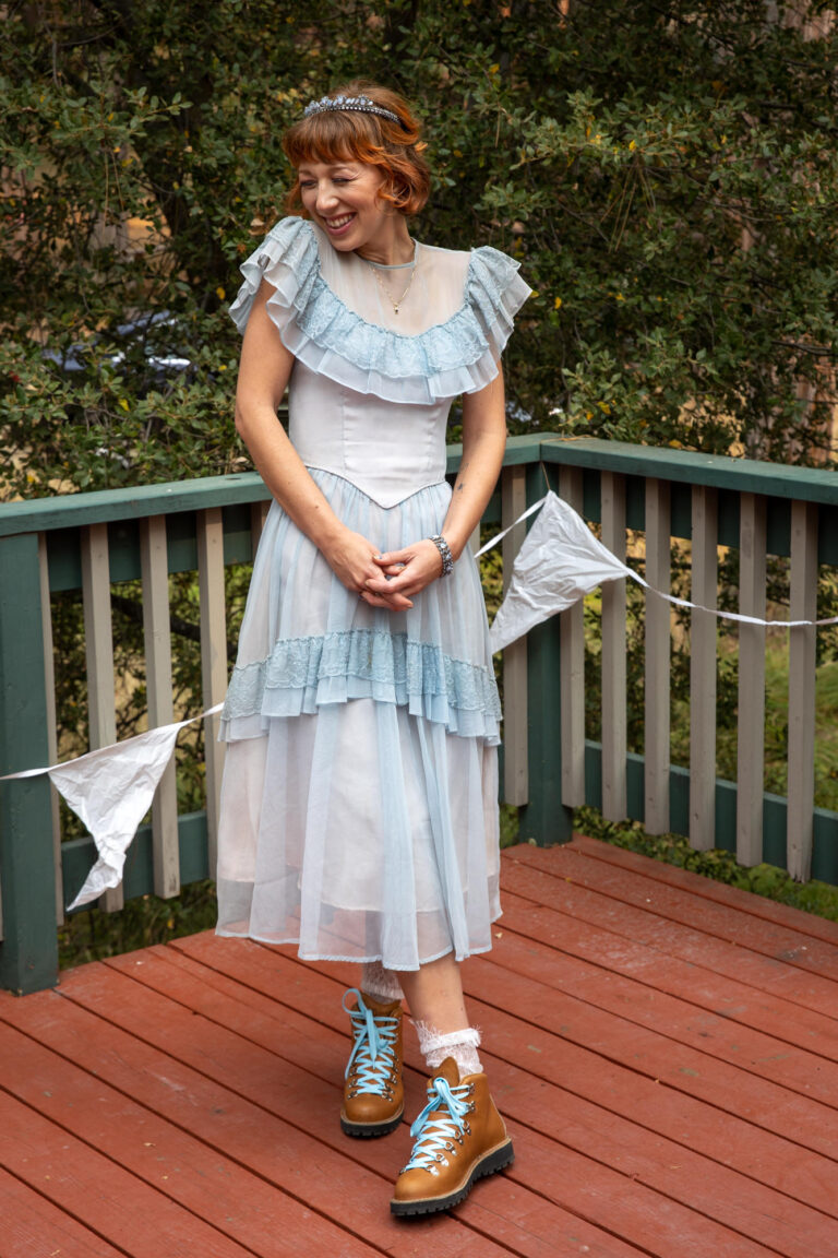 A bride stands with her legs crossed, clasping her hands together and smiling in her blue vintage wedding dress.