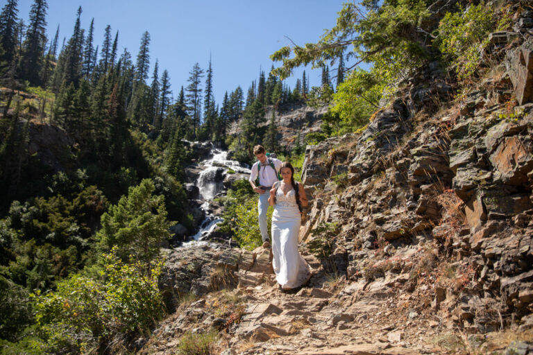 A bride and groom walk down a rocky path with a waterfall behind them.