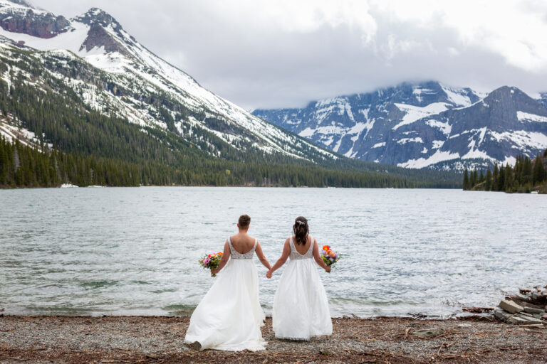 Two brides stand holding hands on the shore of a lake in Montana while holding bouquets in their opposite hands.