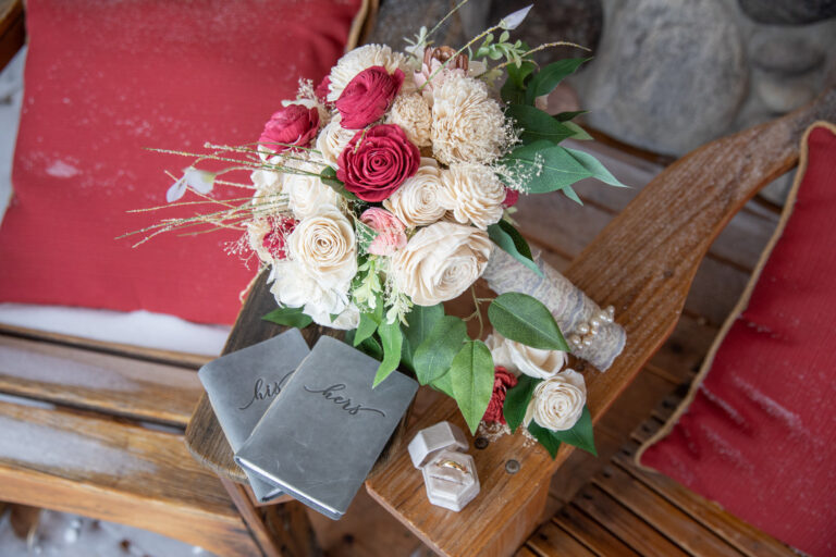 A wedding bouquet, vow books and wedding rings rest on the arm of a wooden chair.