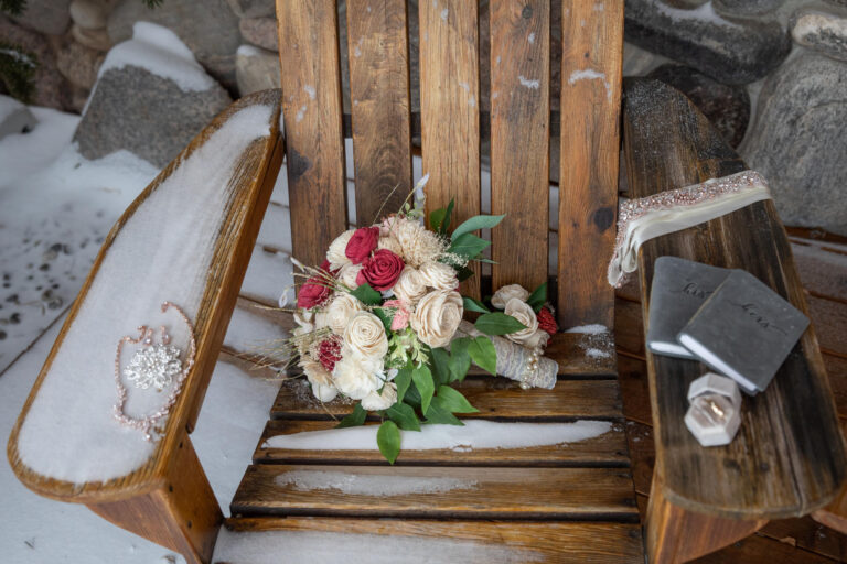 A bouquet of flowers rest on the seat of a wooden chair and a necklace, wedding rings, and vow books rest on the arms of the chair.