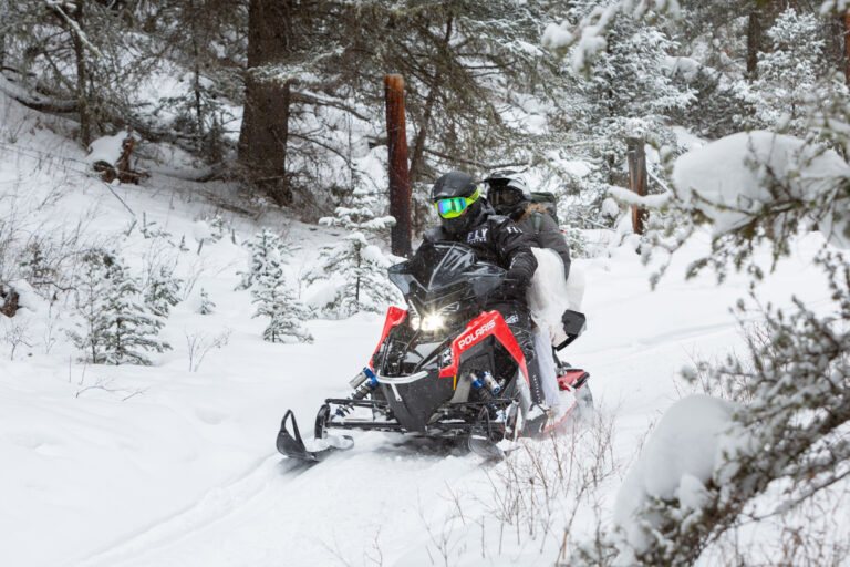 A bride and groom ride on a snowmobile through a forest of trees.