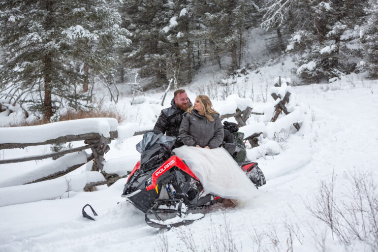 A bride sits on a snowmobile and looks over her shoulder at her groom standing behind her.