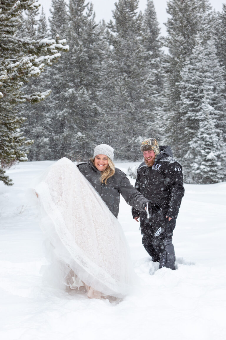 A bride throws her dress up in the air as she walks through the snow and her groom stands behind her laughing.