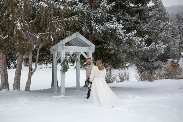 A groom standing near an arch outside in the snow, turns to look at his bride for their first look.