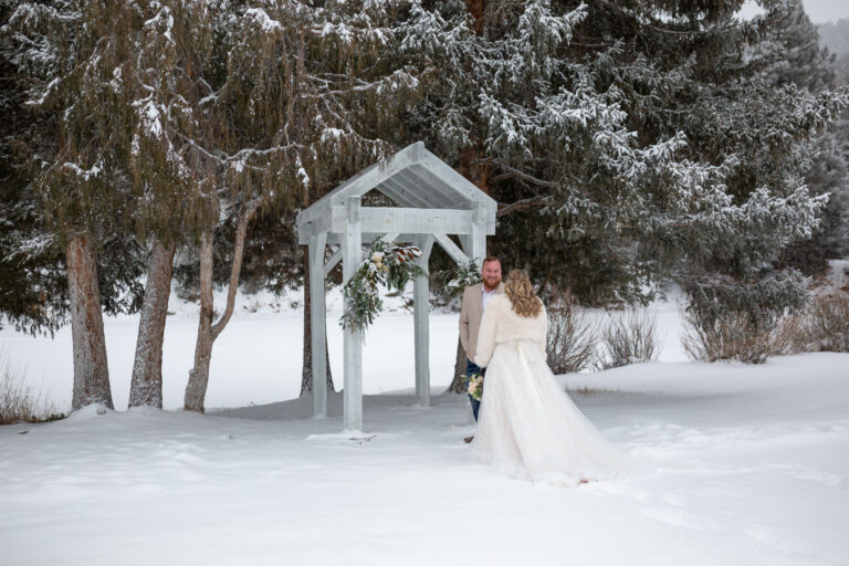 A groom stands in front of an arch while looking at his bride outside in the snow.
