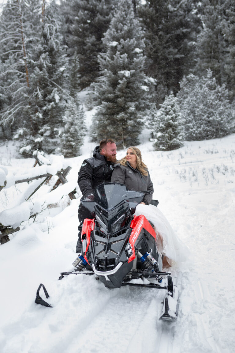 A bride sits on a snowmobile smiling over her shoulder at her groom standing behind her.