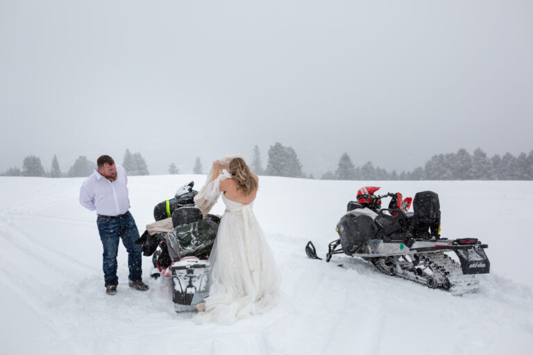 A bride and groom stand next to a snowmobile getting dressed for their winter elopement ceremony.
