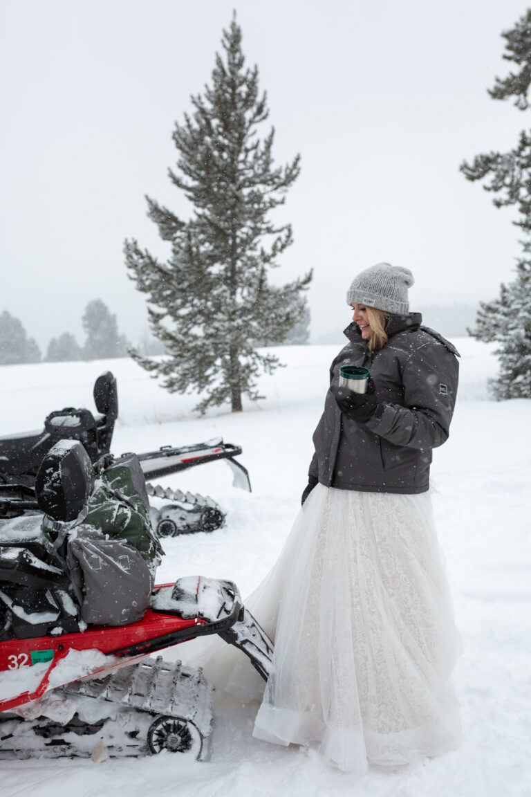 A bride stands behind a snowmobile, holding a cup of hot chocolate.