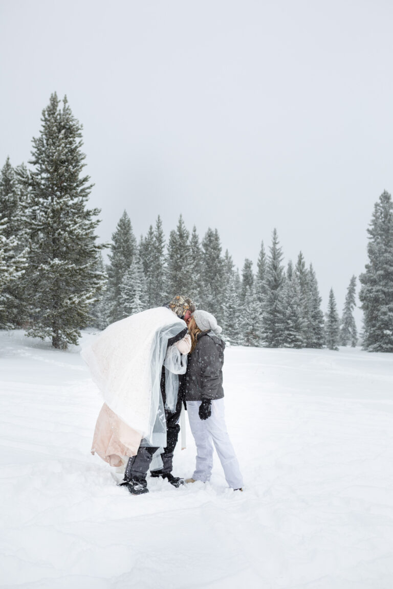 A bride and groom stand in the snow kissing as the groom has the brides wedding dress slung over his shoulder.