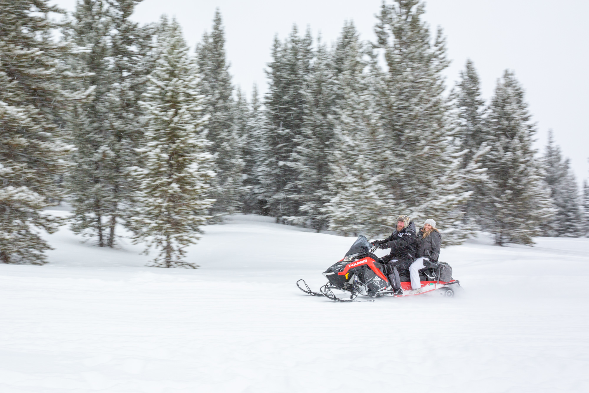 A couple ride on a snowmobile with trees behind them on their winter elopement day.