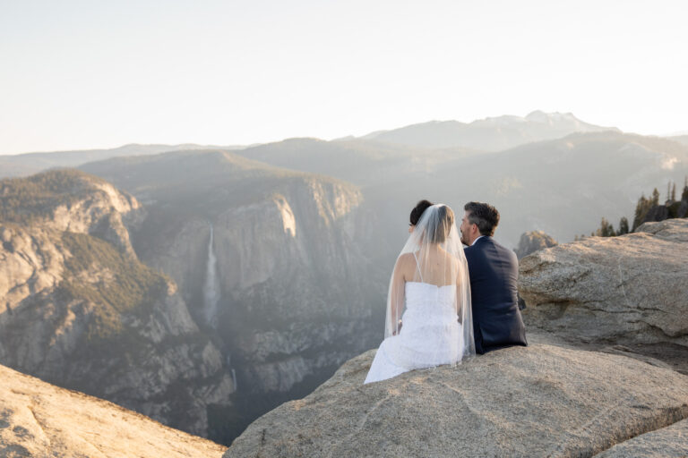 A bride and groom sit on a rock at Taft Point, looking at Yosemite Falls across the valley.