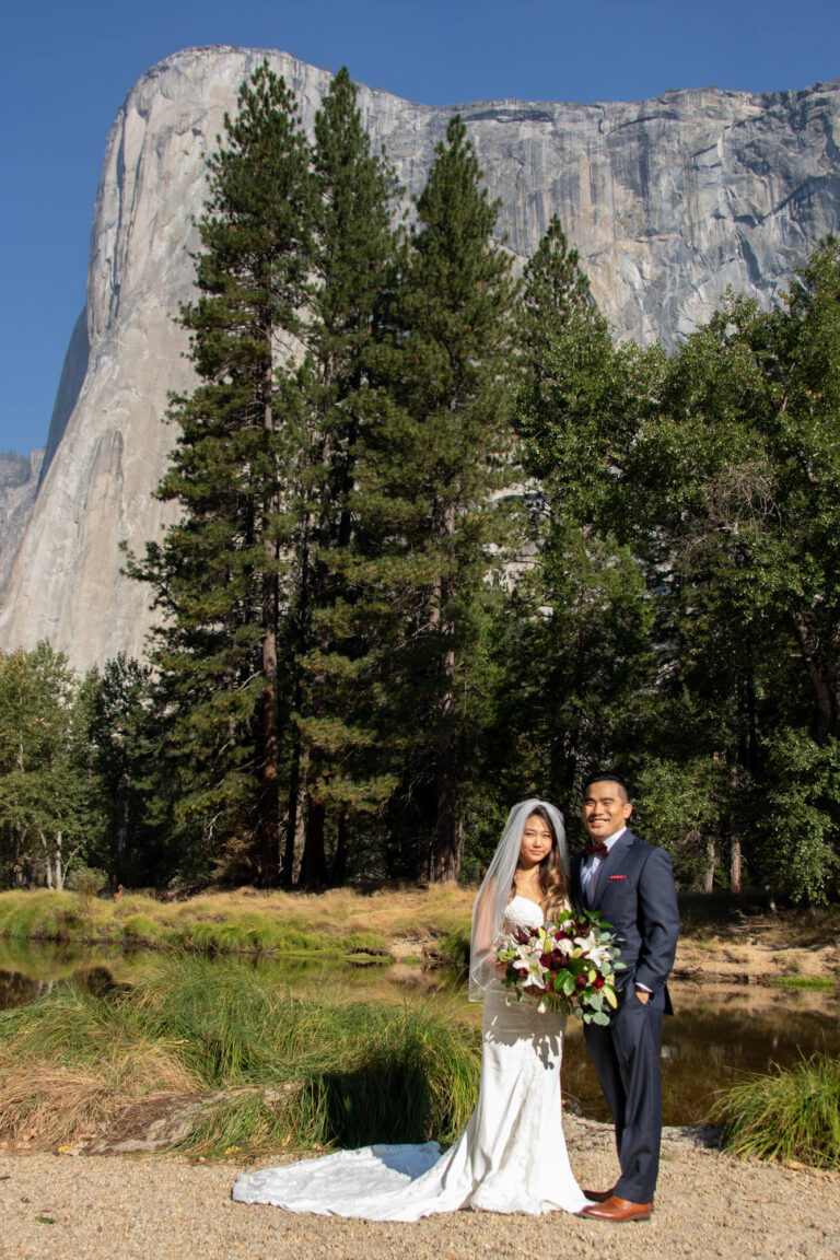 A bride and groom stand smiling in front of El Capitan on a beach on their Yosemite elopement day.