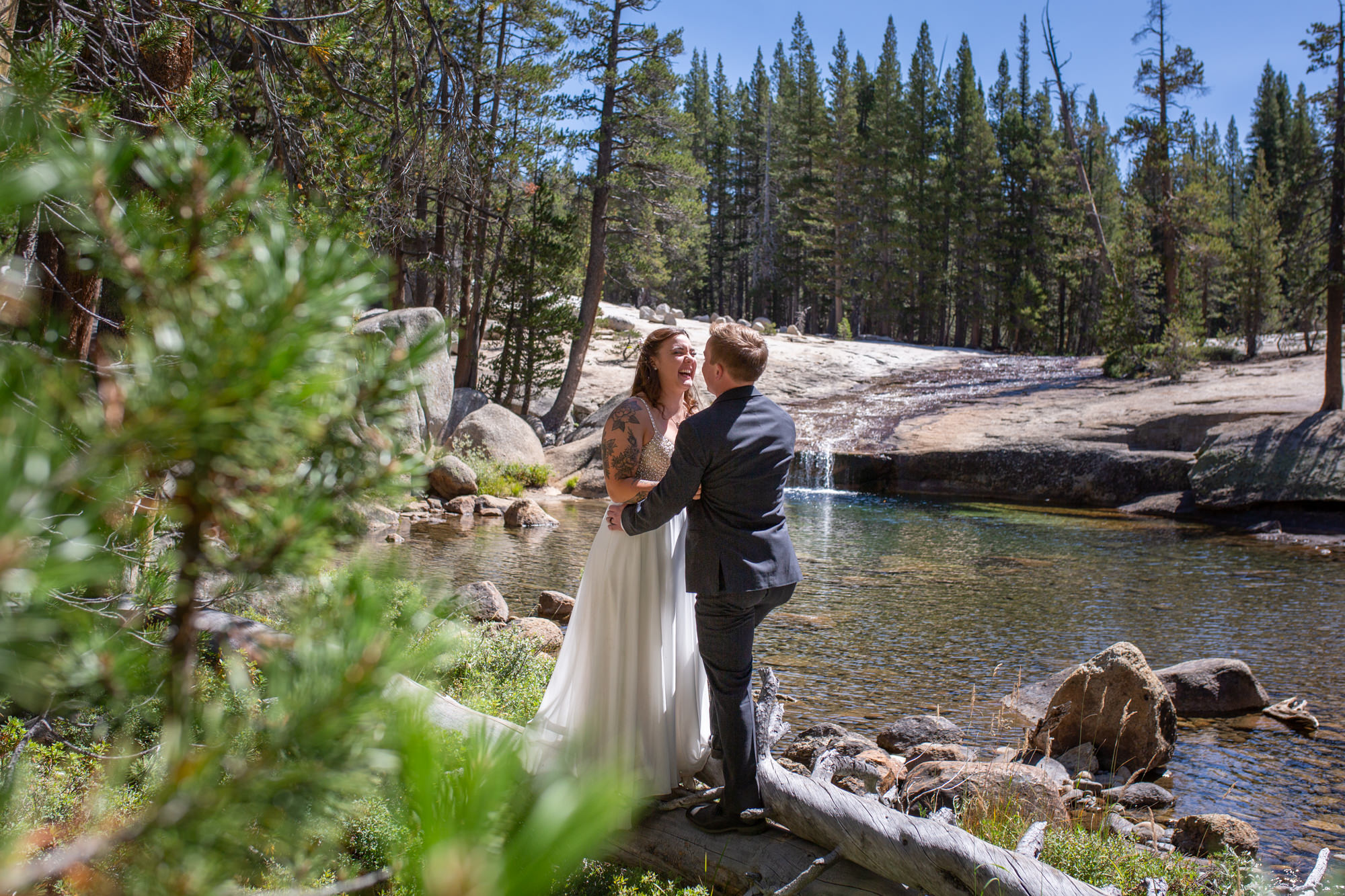 A bride and groom stand on a log next to a river on their Yosemite elopement day.