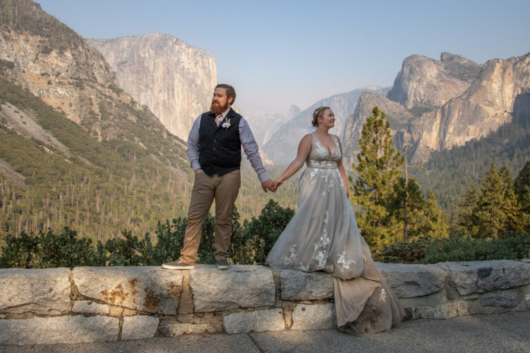 A bride and groom hold hands and stand on a brick wall on their Yosemite elopement day.