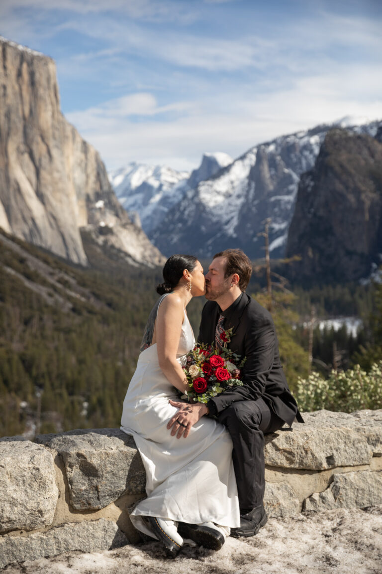 A bride and groom kiss while sitting on a rock wall in Yosemite